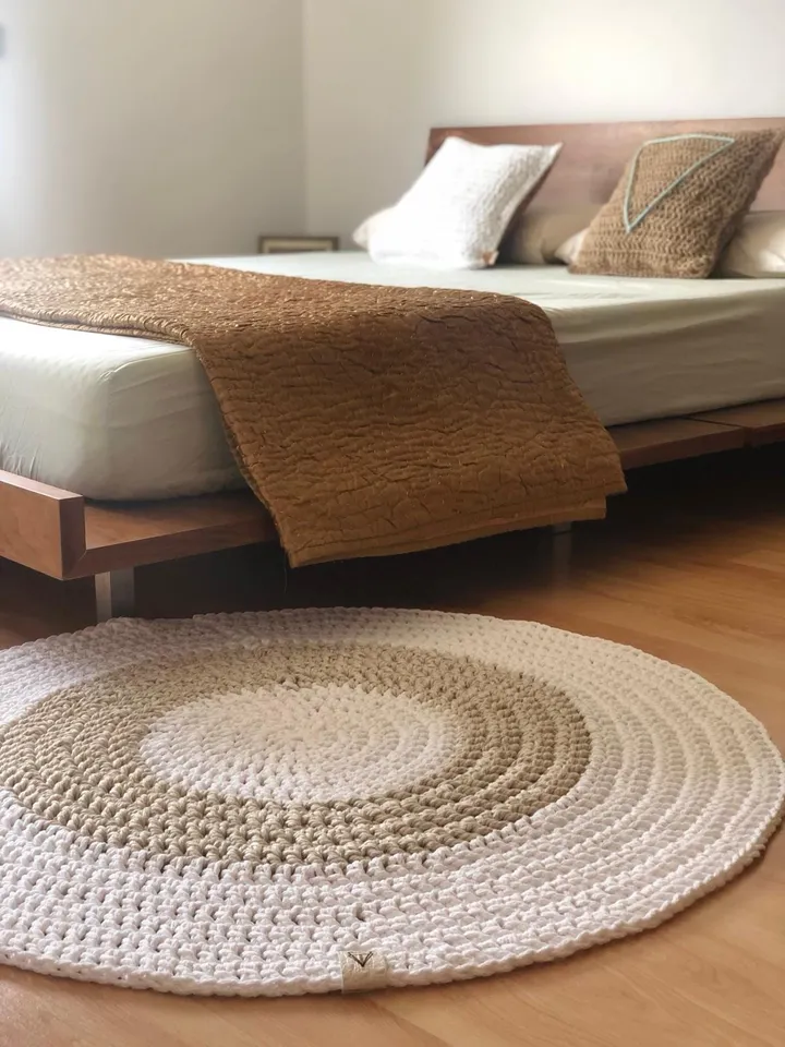 Una alfombra redonda de crochet a los pies de la cama, en un dormitorio, para ver la medida