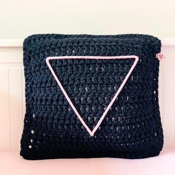 Cojín de crochet en negro con el triángulo de BY S’OHO en cordón rosa, sobre un banco blanco