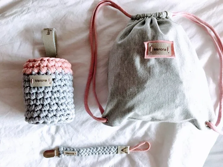 Pack baby de crochet en gris y rosa con el nombre Mariona: portabibis, chupetero y mochilita, en plano