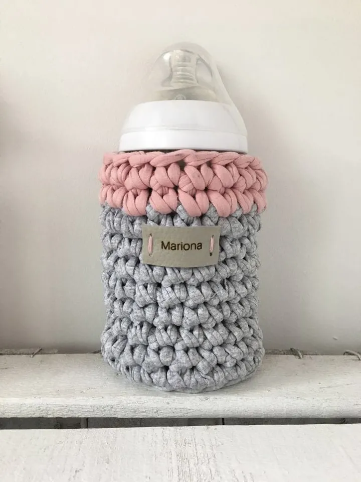 Portabibis de crochet en gris y rosa con el biberón dentro y la etiqueta con el nombre Mariona